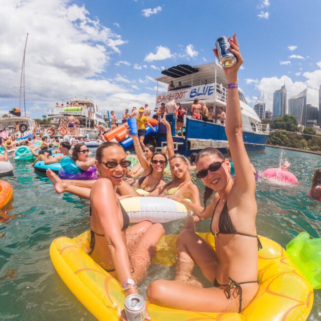 A group of women in swimsuits smile and raise drinks while sitting on floaties in the water at a lively boat party, with many people and boats in the background on a sunny day.