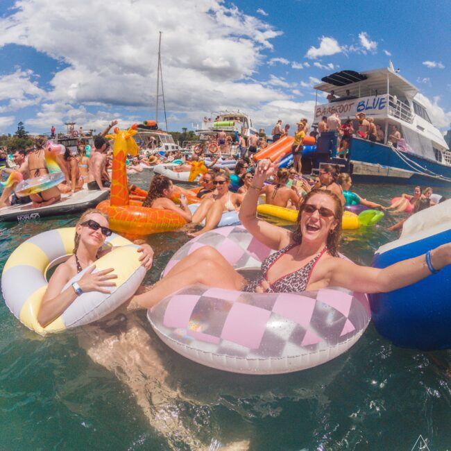 Two women in inflatable pool rings smile and pose for the camera at a lively floating party with many people, colorful floaties, and boats in the background on a sunny day.