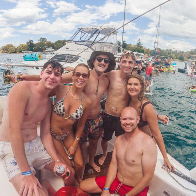 Six smiling young adults in swimwear pose together on a boat, holding drinks, with water and other boats in the background on a sunny day. It appears to be a lively, social gathering.