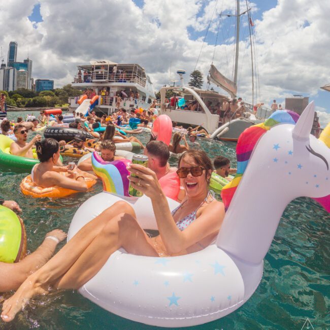 A woman in sunglasses smiles and relaxes on a unicorn pool float, holding a drink, surrounded by people on colorful inflatables in the water near boats, with city buildings and blue sky in the background.