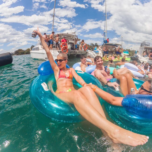 A group of young adults enjoy a sunny day, floating on inflatable tubes in the water, with boats anchored nearby. A smiling woman in sunglasses raises her arm while others relax and hold drinks under a blue sky with scattered clouds.