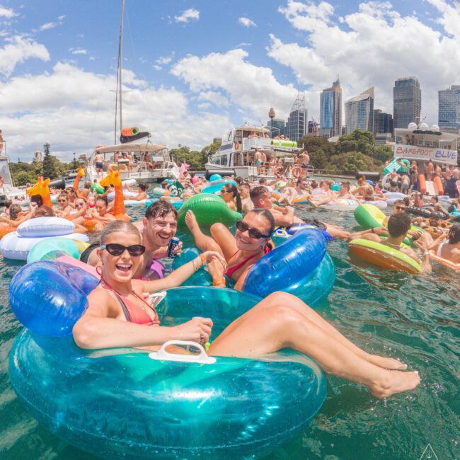 A group of people in swimsuits relax on colorful inflatable tubes and floaties in the water at a lively boat party, with city skyscrapers, boats, and a partly cloudy sky in the background.