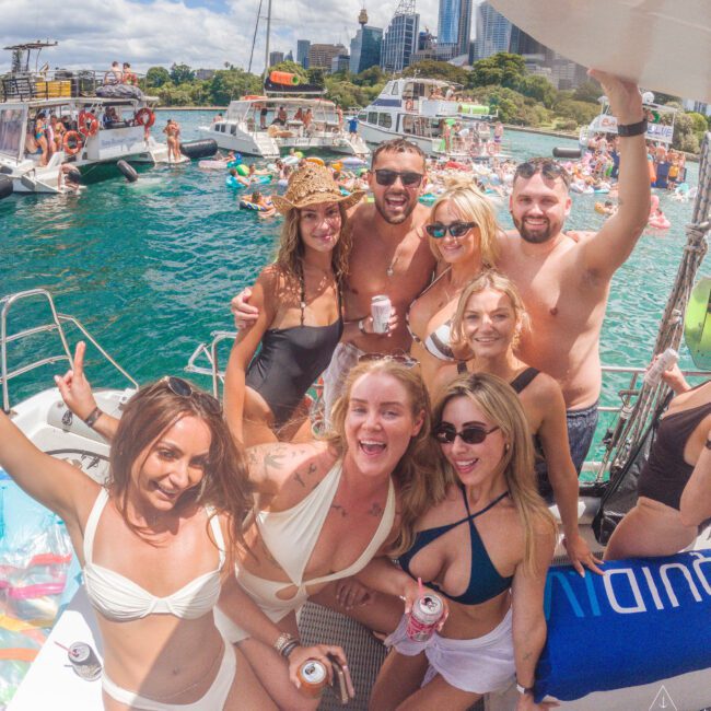 A group of smiling friends in swimsuits pose for a photo on a boat during a sunny day, with other boats, people, and city buildings visible in the background. They appear to be enjoying a lively boat party.