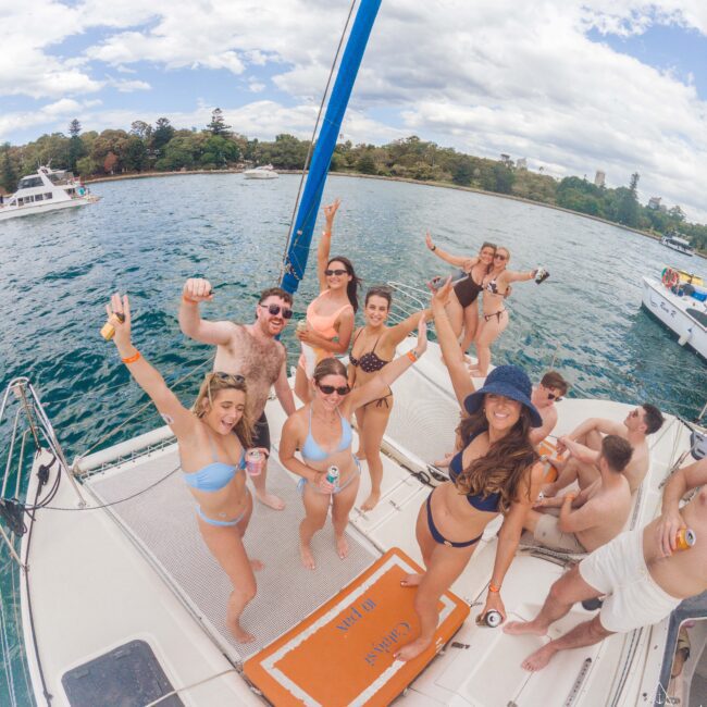 A group of people in swimsuits smile and raise their arms while enjoying drinks on the deck of a sailboat, anchored on a sunny day near a shore with trees and other boats in the background.