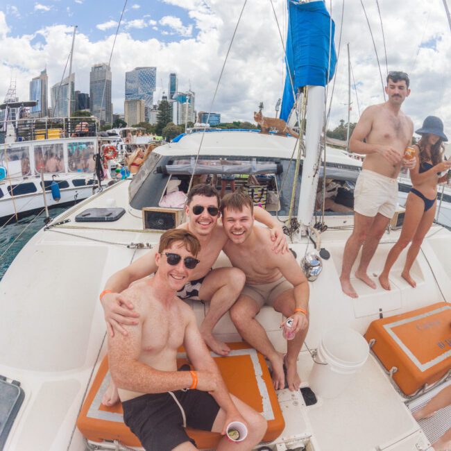 Three young men in swimwear sit and laugh together on a catamaran, holding drinks. Other people relax nearby. The boat is docked by a city skyline under a partly cloudy sky.