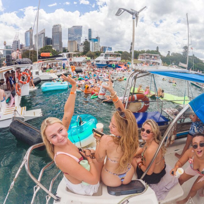 A group of young women in swimsuits smile and pose on a boat, surrounded by other boats and people on colorful inflatables, enjoying a lively summer party on the water with a city skyline in the background.
