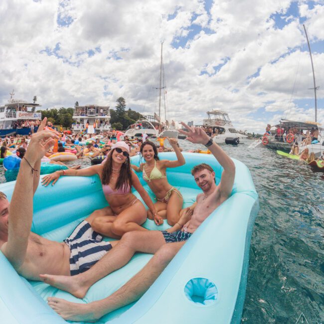 Four young adults in swimsuits relax and smile on a blue inflatable float in the water, surrounded by many boats and other floats under a partly cloudy sky, enjoying a lively outdoor event.
