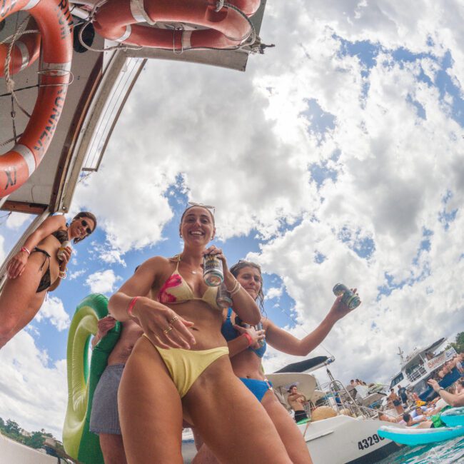 A group of women in swimsuits, smiling and holding drinks, stand on the deck of a boat with lifebuoys, with a bright sky and other boats visible in the background. The scene captures a lively summer atmosphere.