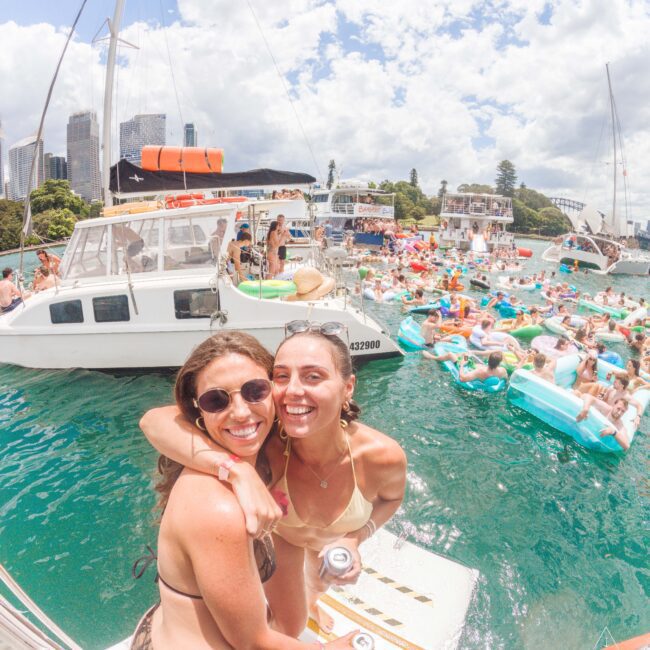 Two smiling women in swimsuits pose on a boat in sunny weather, surrounded by other boats and many people relaxing on inflatable floats in the water. A city skyline and trees are visible in the background.