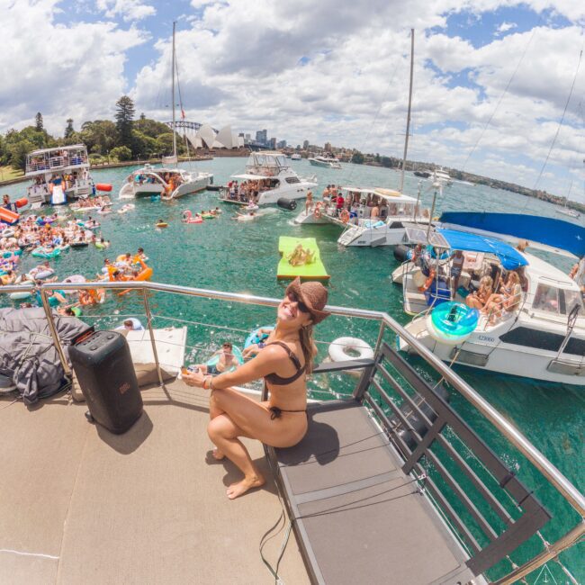 A woman in a black bikini and cap smiles on a boat deck, surrounded by anchored boats and people swimming and relaxing on inflatables in turquoise water under a partly cloudy sky.