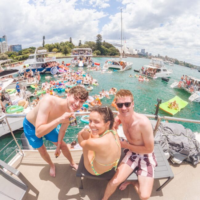 Three young adults in swimwear smile at the camera on a boat, surrounded by other boats and people enjoying a lively party on the water under a partly cloudy sky. The scene is festive and vibrant.