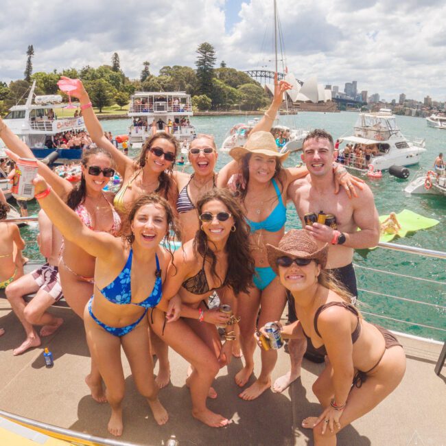 A group of smiling friends in swimwear pose together on a boat, enjoying a sunny day. Other boats and people swimming are visible in the background, with a city skyline and iconic bridge in the distance.