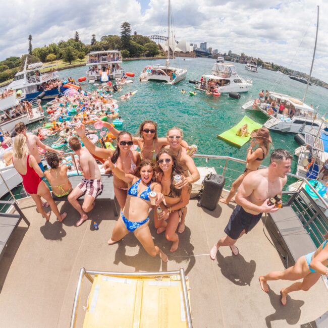 A group of young people in swimsuits pose and smile on a boat, surrounded by others sunbathing and relaxing. Nearby boats and floaties fill the water on a sunny day with a cityscape and trees in the background.
