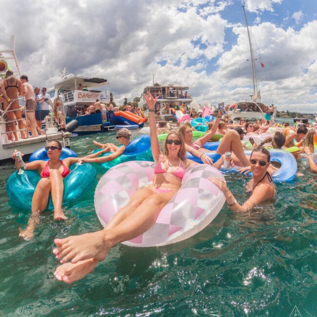 A group of people float on colorful inflatables in the water near several boats under a partly cloudy sky, enjoying a lively party atmosphere. Some hold drinks and smile at the camera.