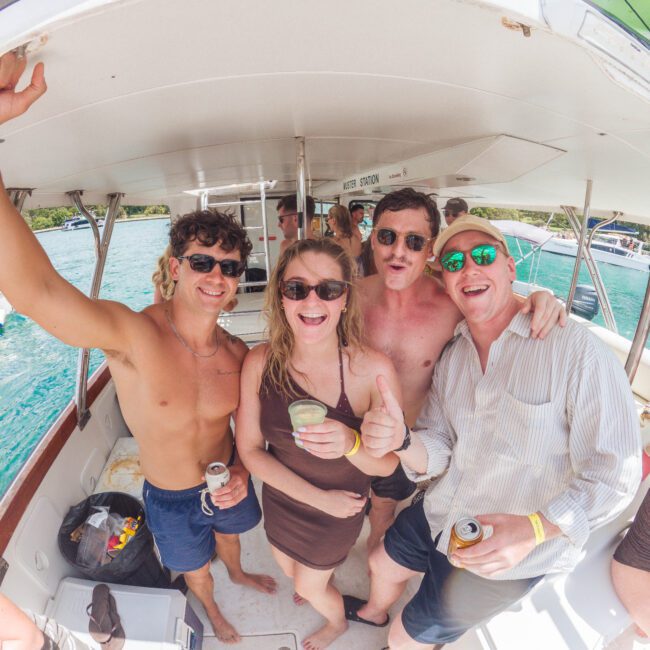 Four smiling people on a boat, two shirtless men in sunglasses, a woman in a brown dress holding a drink, and another man in a striped shirt, all posing closely together near the water on a sunny day.