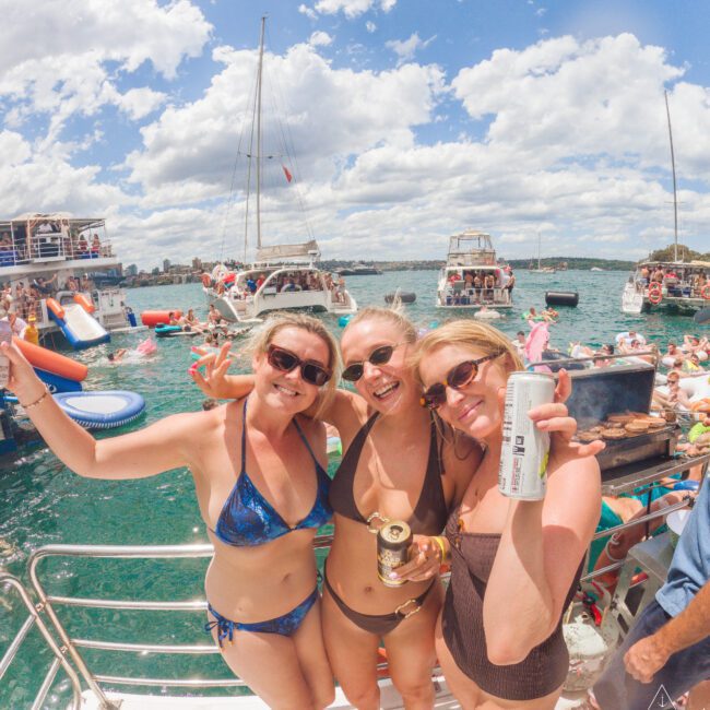 Three women in swimsuits smile and pose with drinks on a boat, surrounded by people, other boats, and floating inflatables on a sunny day with blue sky and scattered clouds.