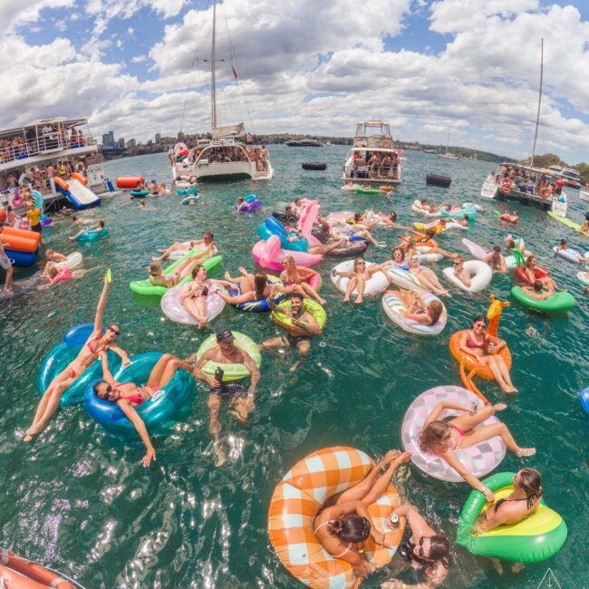 A group of people relax on colorful inflatable floats in the water near boats under a partly cloudy sky, enjoying a lively social gathering.