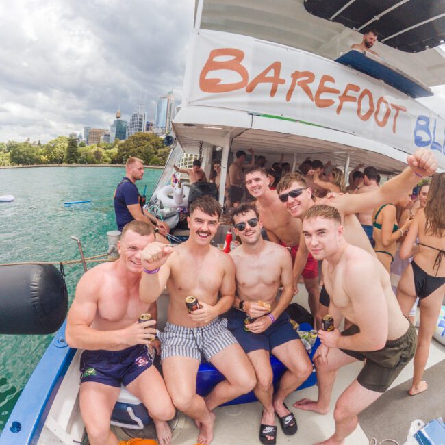 A group of young men in swimwear poses and smiles for the camera on a boat labeled "Barefoot," enjoying drinks, with city buildings and trees visible in the background.