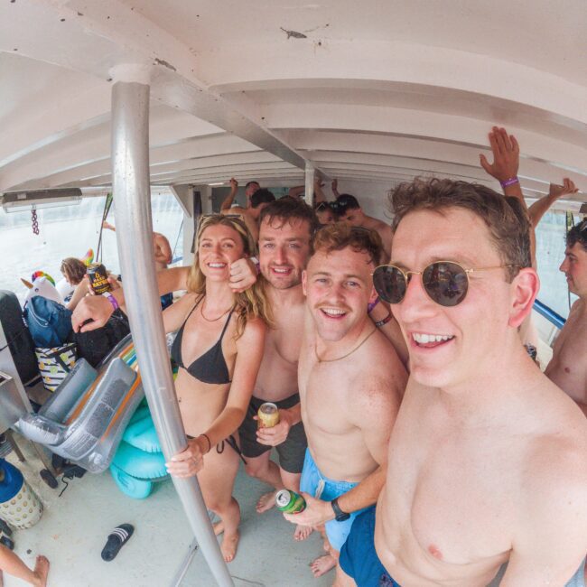 A group of young adults in swimwear smile and pose for a selfie on a boat. The atmosphere is lively, with more people in the background enjoying the party. One person is holding a can, and another is wearing sunglasses.