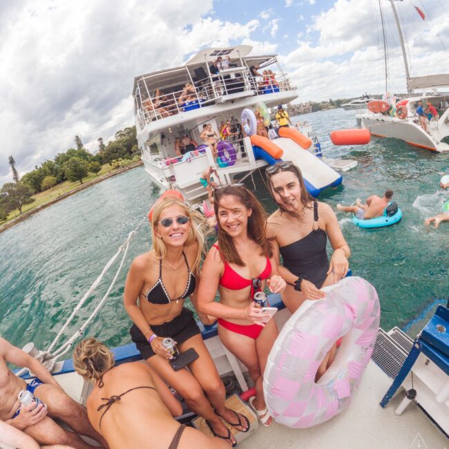 A group of smiling young women in swimsuits pose on a boat, holding drinks and an inflatable pool float. There are other people swimming and relaxing on nearby boats on a sunny day.