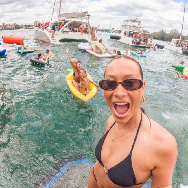 A woman in a black bikini and sunglasses smiles excitedly at the camera in front of party boats and people floating on inflatables in the water on a sunny day.