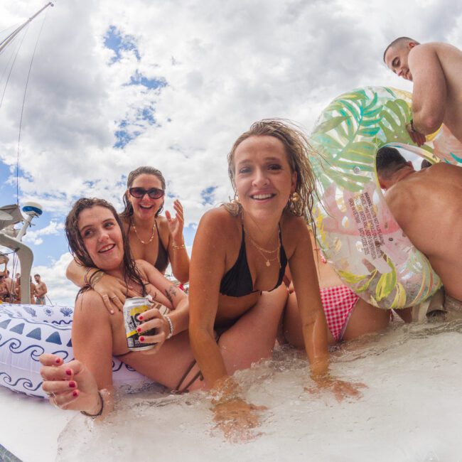 A group of young adults in swimsuits smile and relax together on an inflatable float in the water, enjoying a sunny day with drinks and pool toys. The sky is partly cloudy above them.