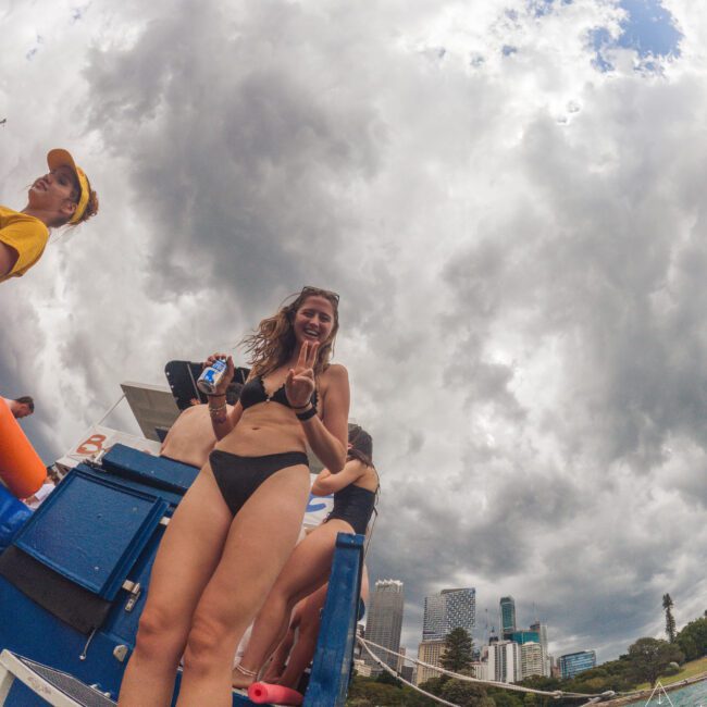 A woman in a black bikini stands on a boat smiling and making a peace sign, holding a drink. Other people are nearby, and city skyscrapers are visible in the background under a cloudy sky.