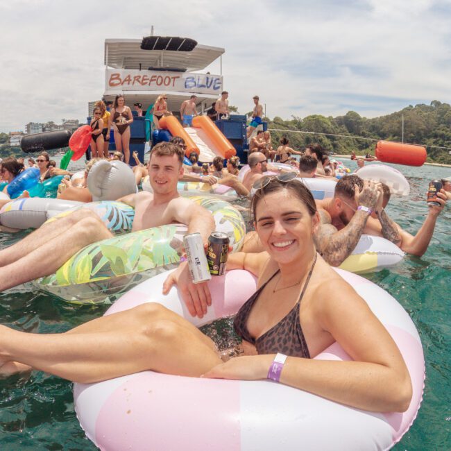 A smiling woman and man relax on inflatable tubes in the water, holding drinks. Other people float nearby, and a boat labeled "Barefoot Blue" is in the background. It's a sunny, festive atmosphere.