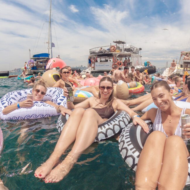 A large group of people relax on colorful inflatable floats in the ocean near boats, smiling and enjoying drinks under a bright, partly cloudy sky. The atmosphere is lively and festive.
