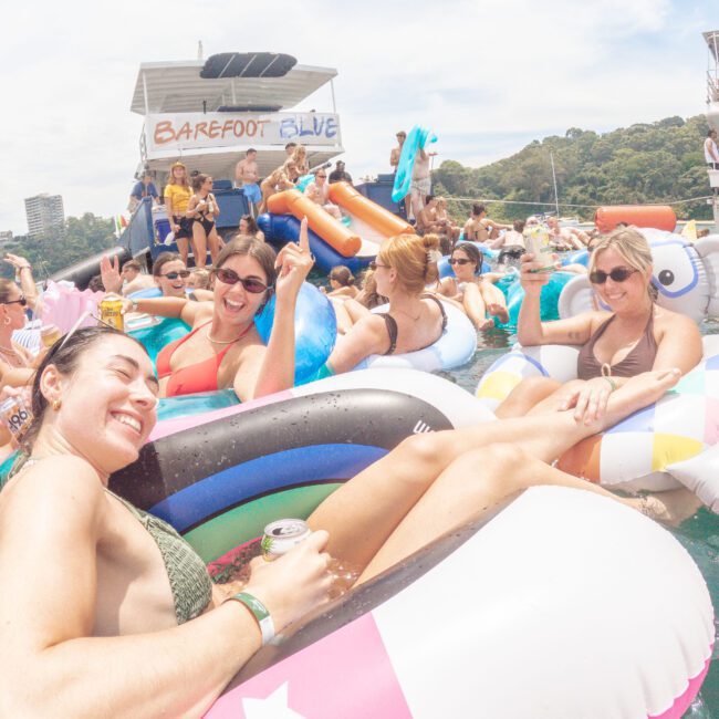 A large group of people in colorful pool floats relax and have fun in the water near boats, enjoying a sunny day. Some hold drinks and smile, with party decorations and an energetic atmosphere all around.