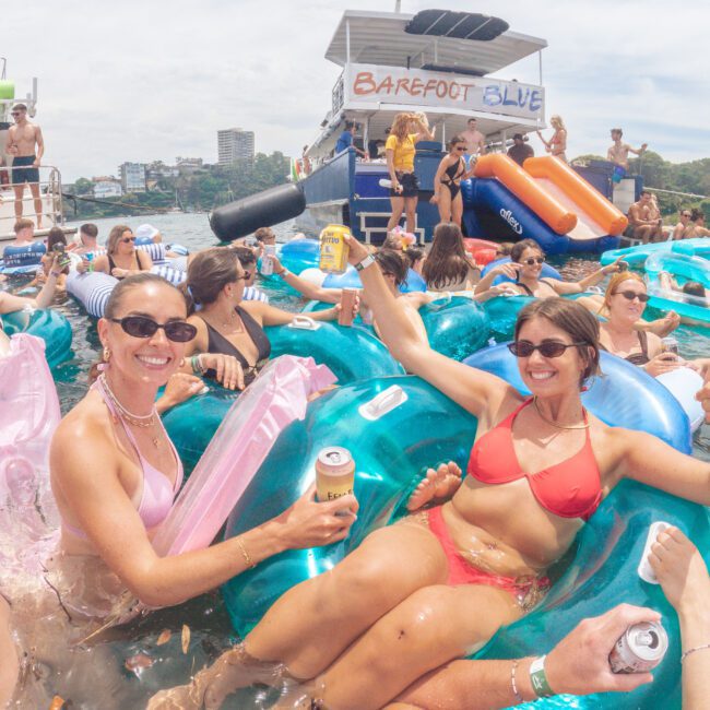 Two women in swimsuits smile and hold drinks while sitting on inflatable floats in a crowded pool. Other people float nearby, and a boat labeled "Barefoot Blue" is visible in the background.