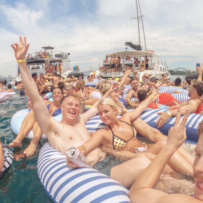 A large group of people relax and smile on striped floaties in the water near anchored boats, enjoying a sunny day. The atmosphere is festive, with drinks in hand and a party vibe.