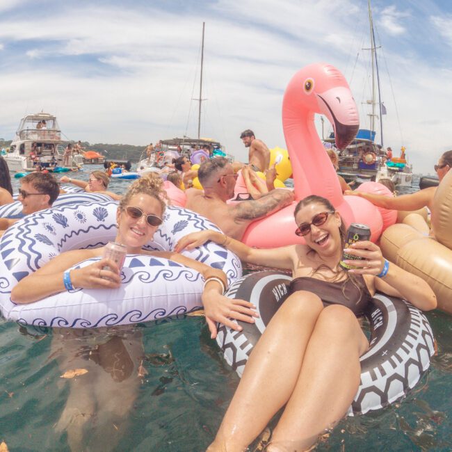 Two women relax on inflatable pool floats, one a flamingo, the other patterned, smiling and holding drinks at a lively yacht party with boats and more people in the background under a blue sky.
