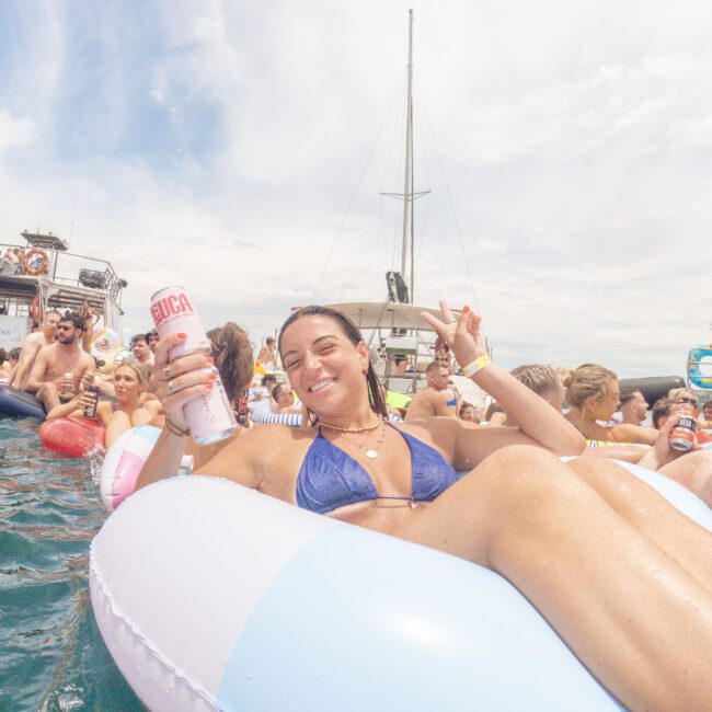 A woman in a blue bikini smiles and holds up a drink can while lounging on a pool float in the ocean, surrounded by other people on floats near boats under a partly cloudy sky.