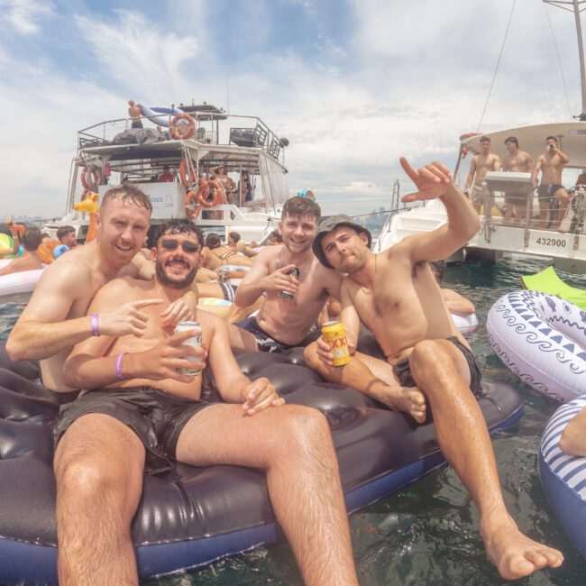Four men in swim trunks smile and pose on a floating mattress in the water, with boats and other people partying in the background under a partly cloudy sky. The atmosphere is festive and relaxed.