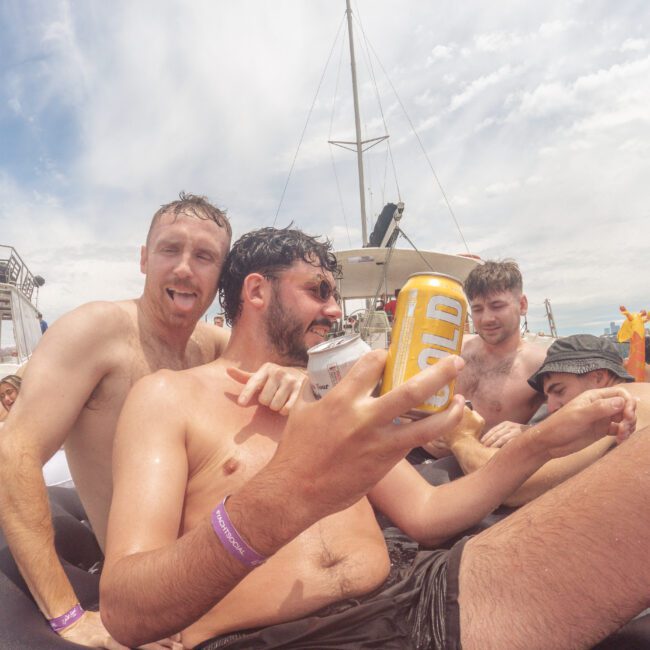 Four young men in swim trunks relax together on an inflatable raft in the water, laughing and holding drinks, with a yacht and cloudy sky in the background. The scene appears lively and carefree.