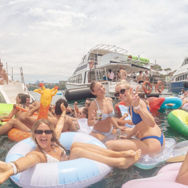 A group of smiling women in swimsuits relax on colorful pool floats in the water, flashing peace signs and holding drinks, with yachts and other people in the background on a sunny day.