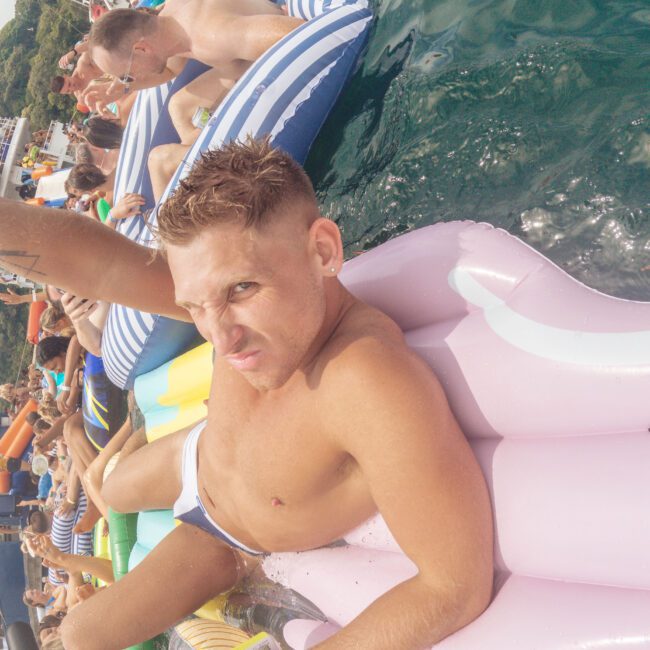 A man lying on a colorful pool float in the water raises his arm and looks at the camera. Many people on floats are around him at what appears to be a lively outdoor pool or lake party.
