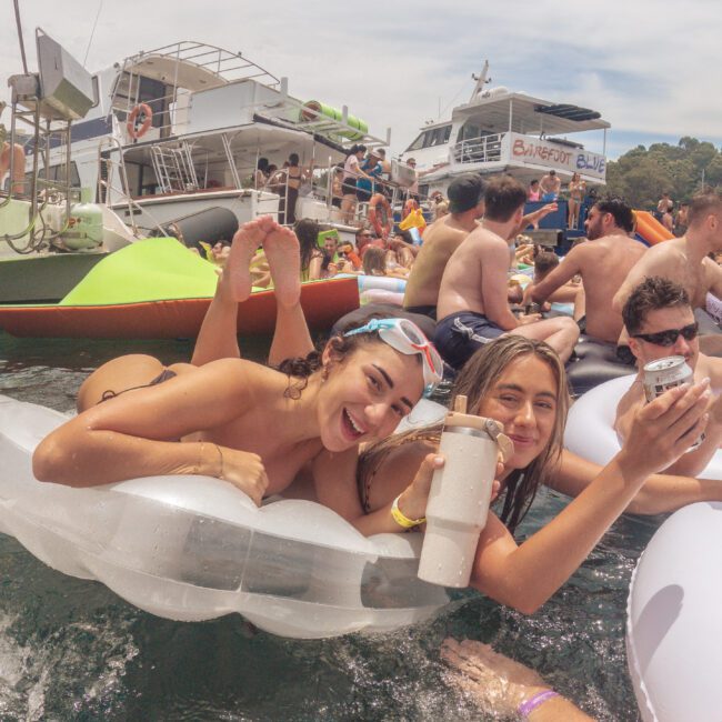 Two young women in swimsuits smile and pose for the camera while floating on an inflatable tube in the water. They are surrounded by other people on floaties and boats, enjoying a lively party atmosphere under a partly cloudy sky.