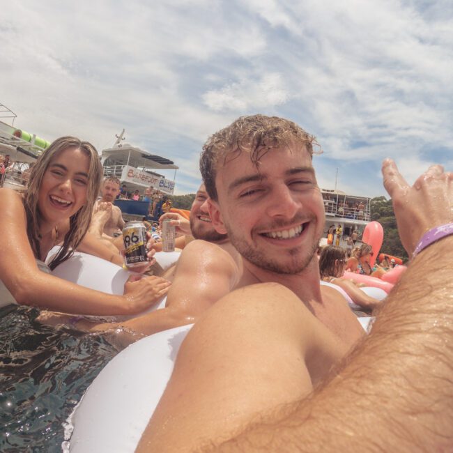 A group of young adults smile and pose for a selfie while relaxing on inflatable pool floats in the water at an outdoor event. The atmosphere is lively and sunny, with boats and other people in the background.