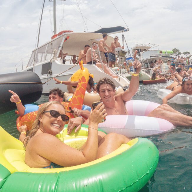 A group of people relax on inflatable pool floats and swim in the ocean near yachts. They smile, hold drinks, and enjoy the sunny day, with boats and more people in the background.