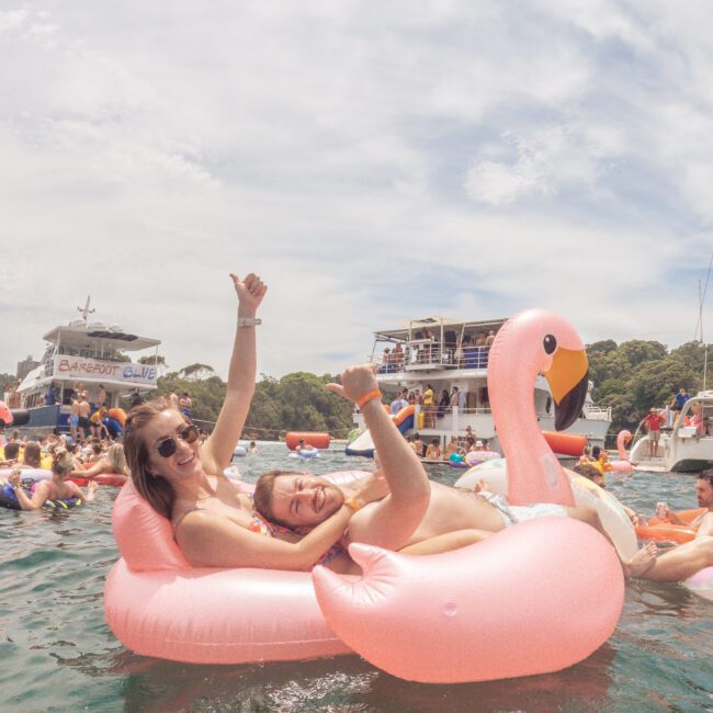 Two people relax and smile on a large pink flamingo float in the water, giving thumbs up. Boats and other people are visible in the background under a partly cloudy sky.