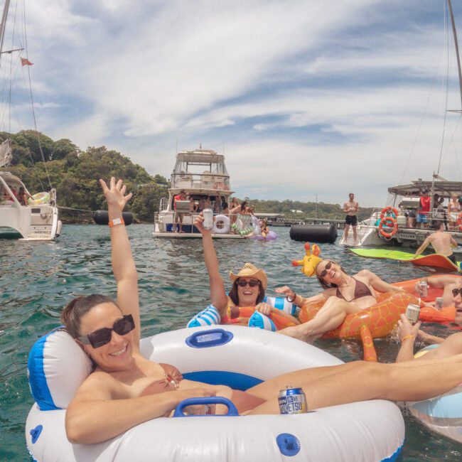 Three women relax on inflatable pool floats in the water, smiling and raising their arms, with boats and other people in the background on a sunny day. The scene is vibrant and festive.