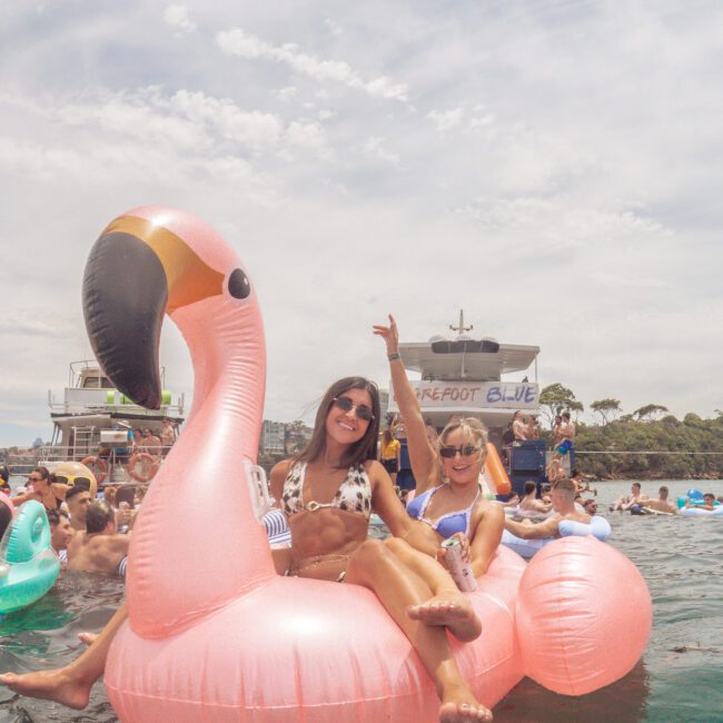 Two women in swimsuits smile and pose on a large pink flamingo pool float in the water, surrounded by other people and boats on a sunny day. One woman holds up a peace sign. The scene looks festive and relaxed.