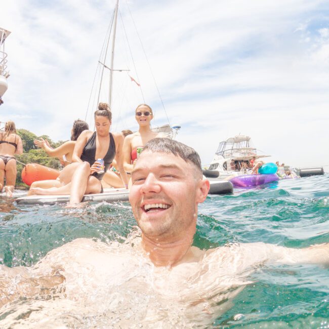 A man smiles in the foreground while swimming in clear blue water. Behind him, several people relax on a floating dock with boats and a sailboat in the background under a partly cloudy sky.