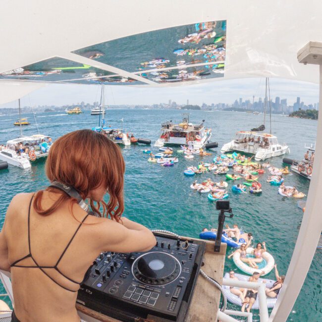A DJ with headphones plays music on a deck overlooking a crowded party on the water, with people on colorful inflatables and boats near a city skyline under a bright sky.