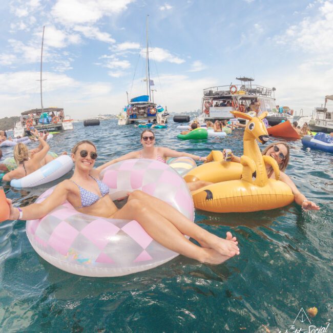 A group of people relax on colorful inflatable pool floats in the ocean near several anchored boats on a sunny day. Three women in the foreground smile at the camera, enjoying the lively social atmosphere.