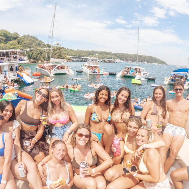 A group of young people in swimsuits smile and pose on a boat with drinks, surrounded by more boats and people swimming and relaxing in a sunny, festive atmosphere on the water.