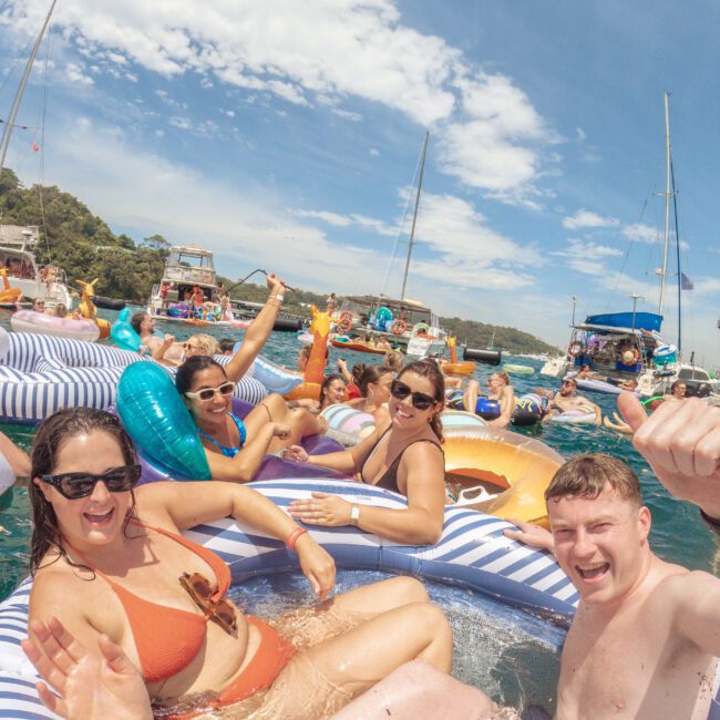 A group of people relax and pose on inflatable pool floats in the water, smiling and waving, with boats and other partygoers in the background under a partly cloudy sky.