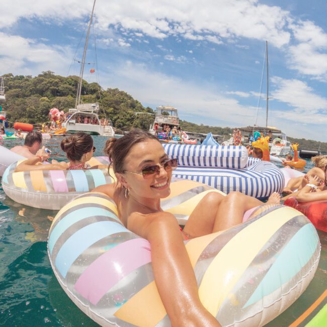 A woman in sunglasses smiles while lounging on a colorful inflatable ring in the water, surrounded by others on floats with boats and greenery in the background under a partly cloudy sky.